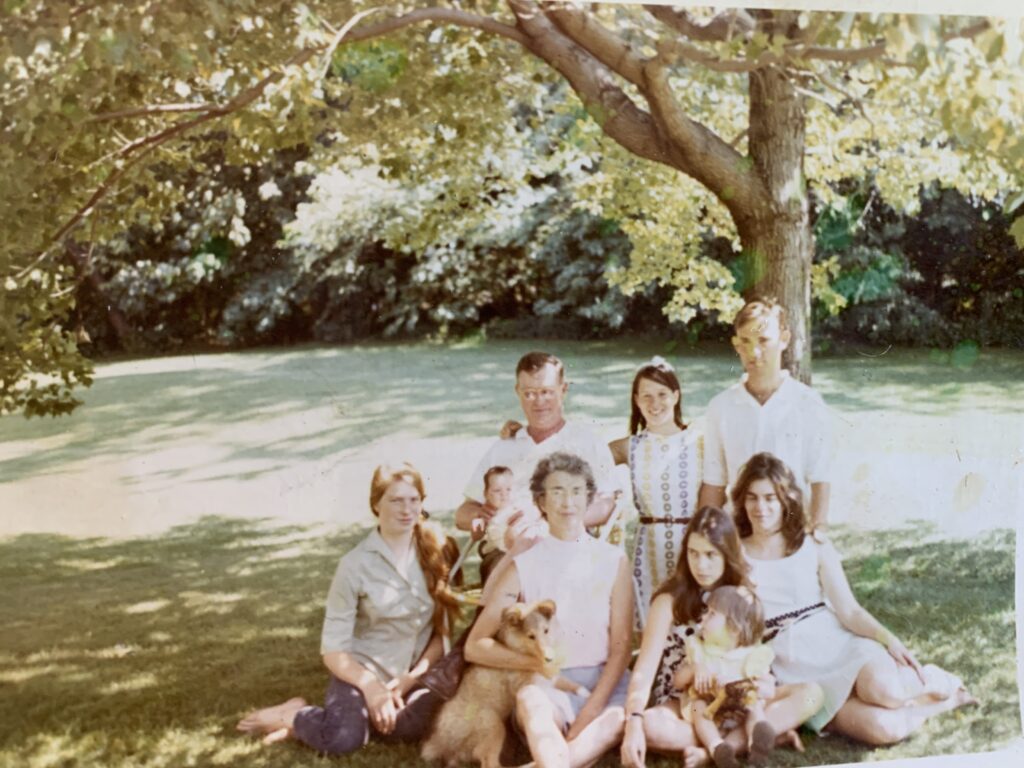A family of nine, three generations seated outdoors in summertime under a tree. The author is two years old, sitting on a young teenage girl's lap. The author is holding a stuffed lion and looking towards her grandmother, who is sitting ramrod straight up, right next to her.